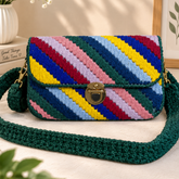 A crocheted shoulder bag with diagonal multicolor stripes in green, blue, yellow, pink, and red, featuring a dark green crocheted strap and a gold-tone clasp, displayed on a neutral background with decorative props.