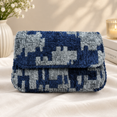 A textured, chunky knit clutch bag with a blue and gray pixelated pattern, resting on a white fabric surface with decorative props in the background.