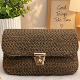 A brown woven clutch bag with a gold clasp, styled on a white faux fur surface with a potted plant and a framed print in the background.