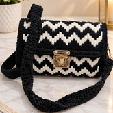 A crocheted handbag with a black and white chevron pattern and a gold clasp, resting on a marble surface with a potted plant and a lantern in the background.