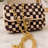 A small checkered flap handbag with a plush bouclé texture in black and beige, featuring a gold-tone chain strap and turn-lock closure, displayed on a marble surface.