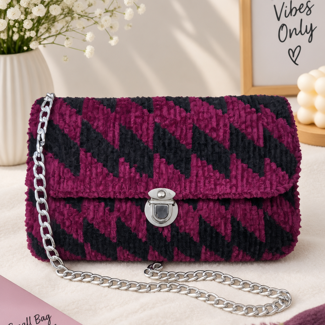 A textured chevron-patterned handbag in deep magenta and black, featuring a silver chain strap and a silver clasp, styled on a cream fabric surface with decorative props including a pink book, white flowers, and a framed sign.