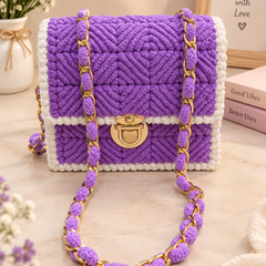 A purple and white crocheted handbag with a gold clasp and matching purple-and-gold chain strap, styled on a tabletop with flowers, a framed sign, and books in the background.