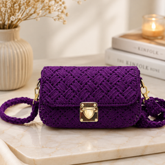 A purple crocheted clutch bag with a gold clasp, resting on a marble tray with dried flowers and books in the background.