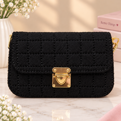 A black quilted textured shoulder bag with a gold-tone padlock clasp, styled on a marble surface with baby's breath flowers and pink books in the background.