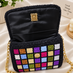 A black fuzzy shoulder bag with a gold chain strap, featuring a colorful checkered pattern on the front flap and a gold clasp, displayed on a beige satin fabric background.