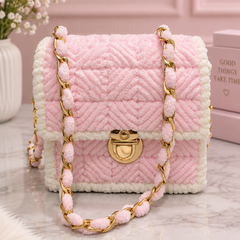 A pink quilted faux-fur handbag with white trim and a gold-tone chain strap, displayed on a marble surface with a pink background and decorative props.