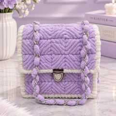 A lavender and white textured quilted shoulder bag with a silver chain strap, styled on a marble surface with a purple vase of flowers and books in the background.