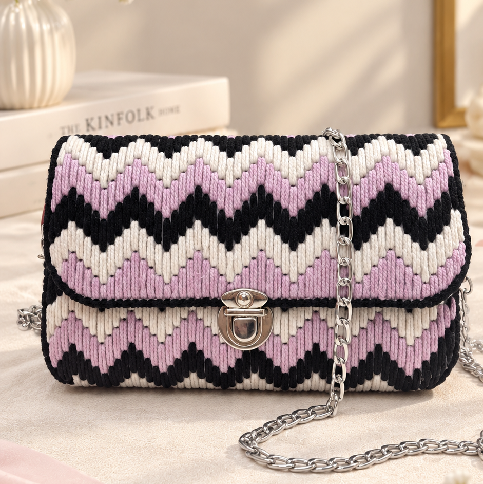 A woven shoulder bag with a black, white, and light pink chevron pattern, featuring a silver-tone chain strap and a silver turn-lock clasp. The bag is styled on a light beige fabric surface with a pink fabric accent, white baby's breath flowers, and a book in the background.