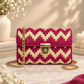 A woven clutch handbag with a magenta and cream chevron pattern and a gold chain strap, styled on a marble surface with decorative flowers and candles.