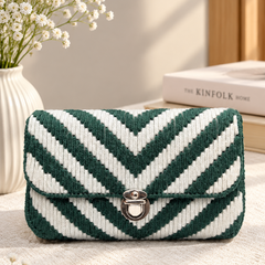 A woven clutch bag with a green and white chevron pattern and a silver turn-lock clasp, styled on a cream fabric surface with baby's breath flowers and a book in the background.