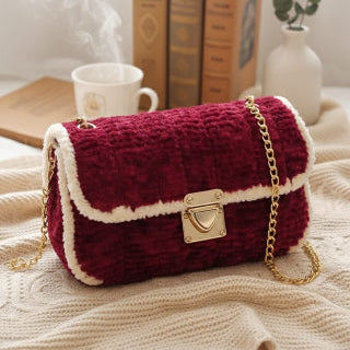 Red textured handbag with gold chain on a beige surface with books and a plant in the background