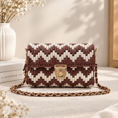 A woven shoulder bag with a brown and cream chevron pattern and a gold-tone chain strap, styled on a light fabric surface with a white vase of dried flowers in the background.