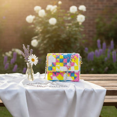 Colorful woven handbag on a table outdoors with flowers and a vase.
