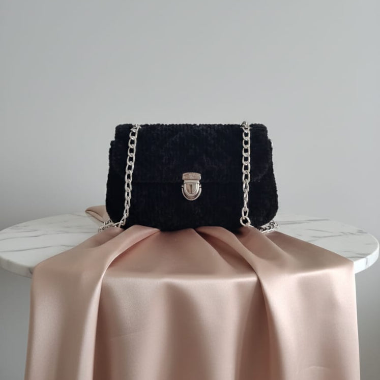 Black textured handbag with a chain strap on a beige fabric surface against a gray background