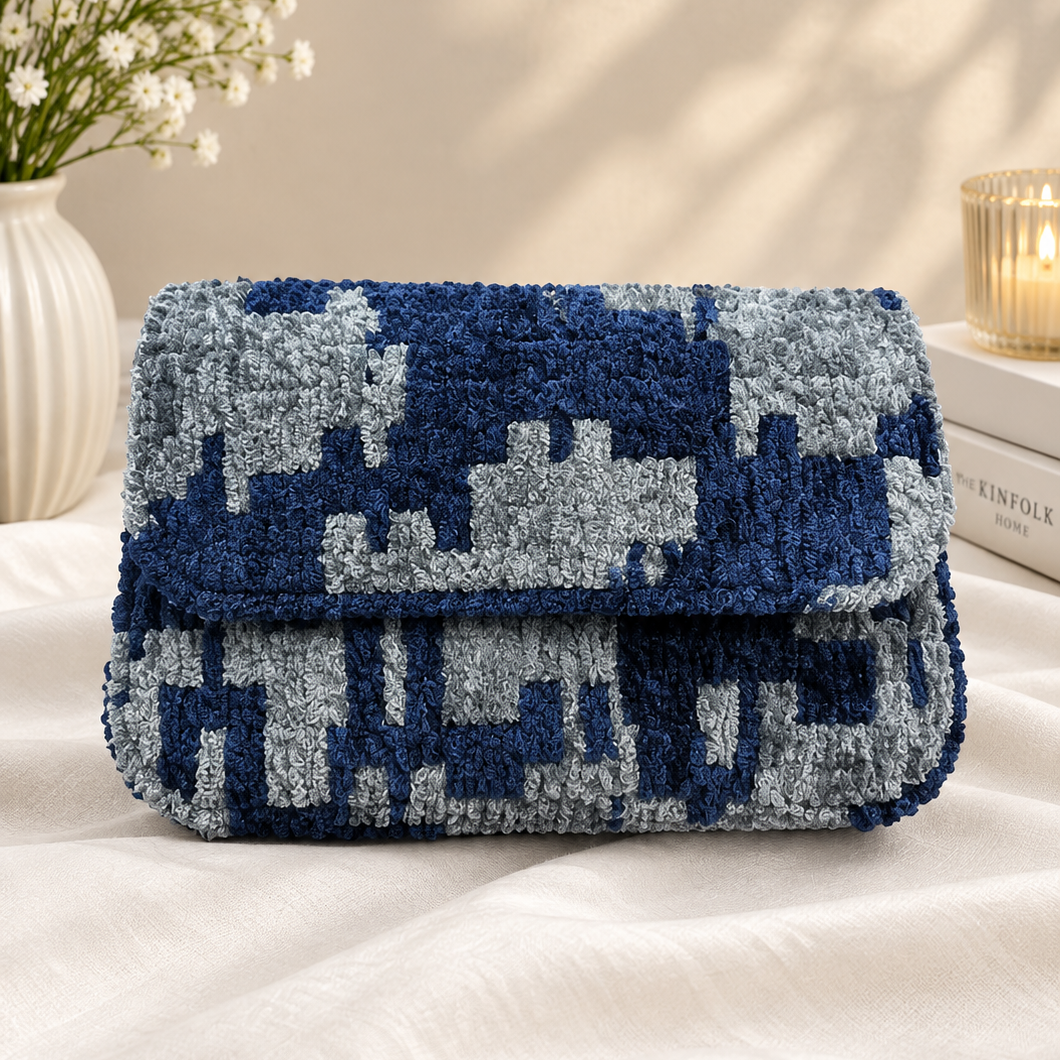A textured, chunky knit clutch bag with a blue and gray pixelated pattern, resting on a white fabric surface with decorative props in the background.
