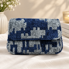 A textured, chunky knit clutch bag with a blue and gray pixelated pattern, resting on a white fabric surface with decorative props in the background.