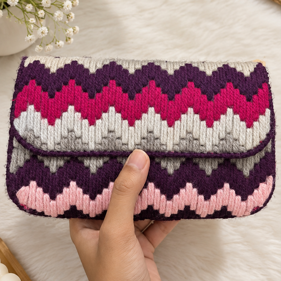 A hand holding a knitted clutch wallet with a chevron pattern in purple, pink, white, and gray, set against a soft white faux fur background with decorative flowers and a woven mat.
