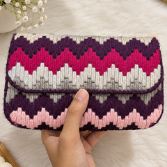 A hand holding a knitted clutch wallet with a chevron pattern in purple, pink, white, and gray, set against a soft white faux fur background with decorative flowers and a woven mat.