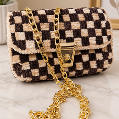 A small checkered flap handbag with a plush bouclé texture in black and beige, featuring a gold-tone chain strap and turn-lock closure, displayed on a marble surface.