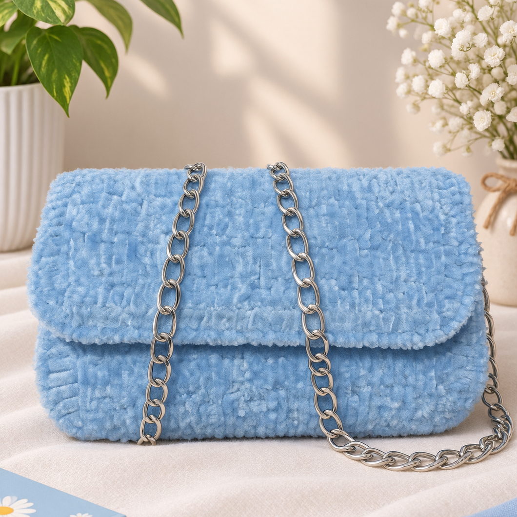 A light blue textured faux fur handbag with a silver chain strap, styled on a beige fabric surface with a potted plant, baby's breath flowers, and a small book in the background.