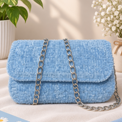 A light blue textured faux fur handbag with a silver chain strap, styled on a beige fabric surface with a potted plant, baby's breath flowers, and a small book in the background.