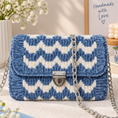 A blue and white chevron-patterned crocheted handbag with a silver chain strap and metal clasp, styled on a light surface with flowers, a book, and a framed sign in the background.