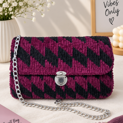 A textured chevron-patterned handbag in deep magenta and black, featuring a silver chain strap and a silver clasp, styled on a cream fabric surface with decorative props including a pink book, white flowers, and a framed sign.
