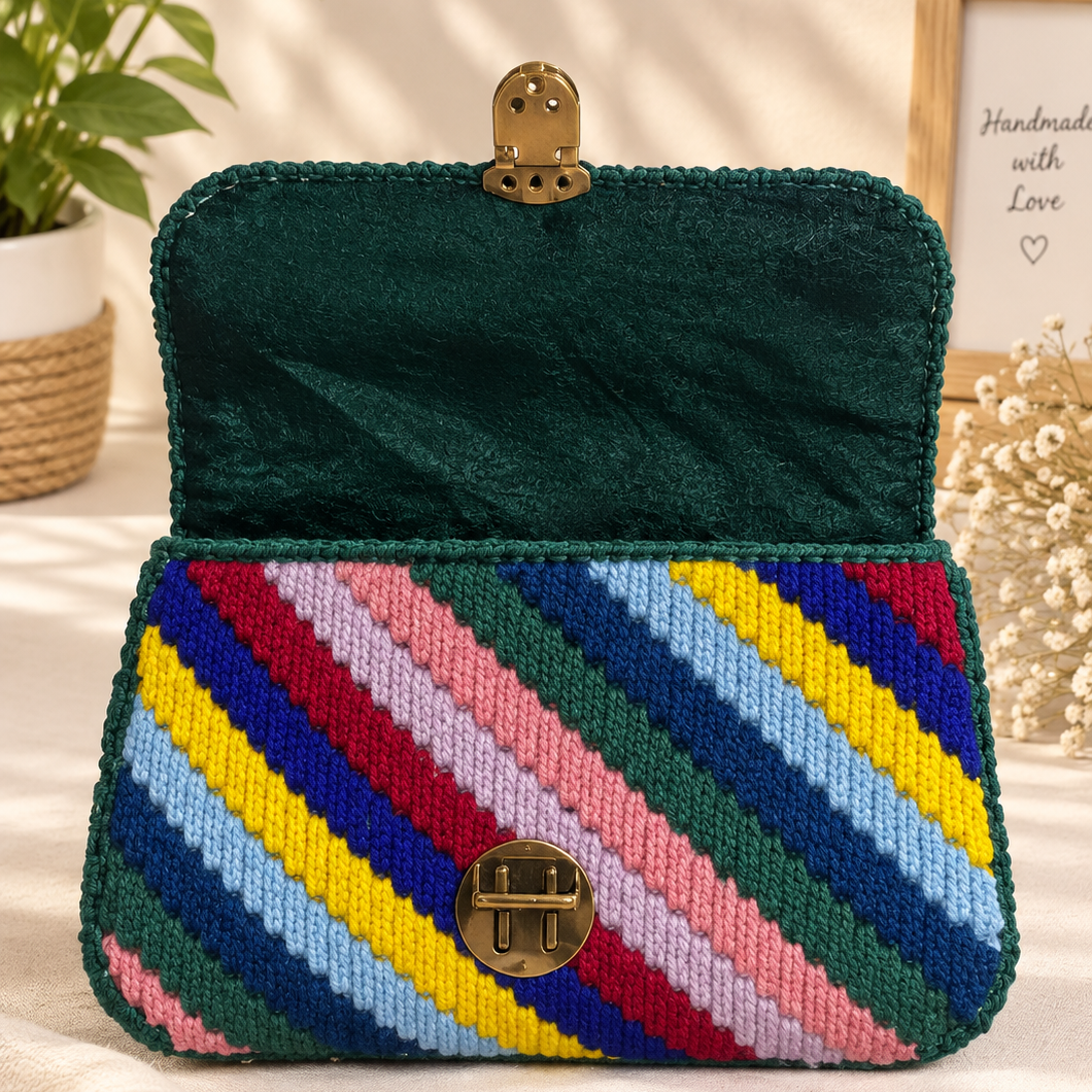 A structured handbag with a dark green velvet interior and flap, featuring a colorful striped woven exterior in shades of blue, yellow, pink, red, and green. It has a gold-tone clasp closure and is styled on a beige fabric surface with decorative plants and a framed print in the background.