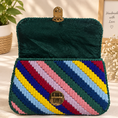 A structured handbag with a dark green velvet interior and flap, featuring a colorful striped woven exterior in shades of blue, yellow, pink, red, and green. It has a gold-tone clasp closure and is styled on a beige fabric surface with decorative plants and a framed print in the background.