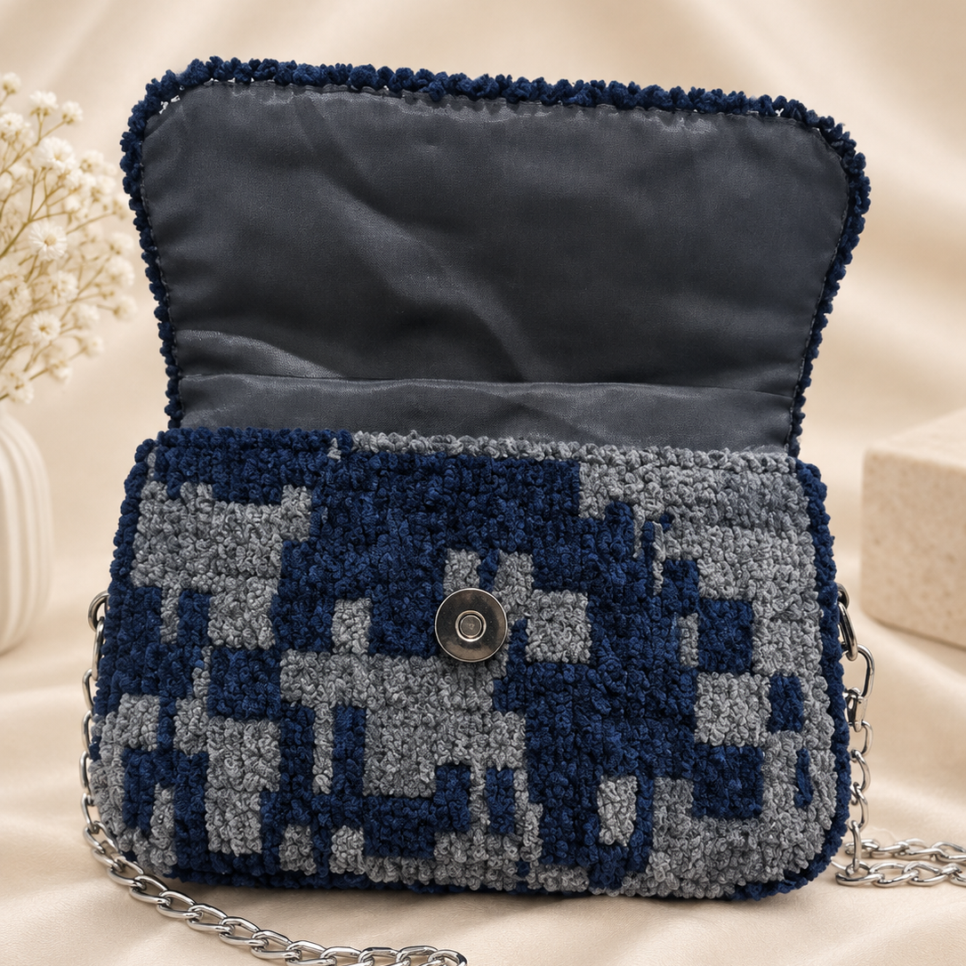 A textured bouclé shoulder bag in navy blue and gray with a geometric pattern, featuring an open top flap, a silver-tone snap button, and a silver chain strap, displayed on a beige satin fabric background.