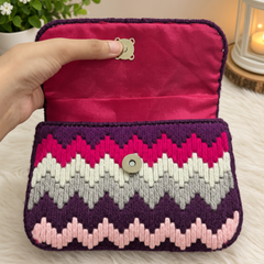 A hand holds open a small woven clutch bag with a magenta velvet interior and a chevron patterned exterior in purple, white, pink, and light purple, set against a soft white faux fur background with a plant and lantern in the background.