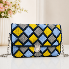  A woven clutch bag with a geometric diamond pattern in yellow, light blue, gray, and navy, featuring a silver turn-lock clasp and a silver chain strap, set against a soft beige background with a blurred floral arrangement.