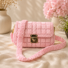 A pink quilted woven shoulder bag with a chunky knotted strap and gold-tone hardware, styled on a beige fabric surface with dried flowers and pink hydrangeas in the background.