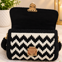 A crocheted shoulder bag with a black and white chevron pattern, featuring a black velvet interior lining and gold-tone hardware, displayed on a white faux fur surface with decorative props.