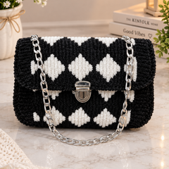 A black and white diamond-patterned crocheted handbag with a silver chain strap and clasp, styled on a marble surface with flowers, books, and a lantern in the background.