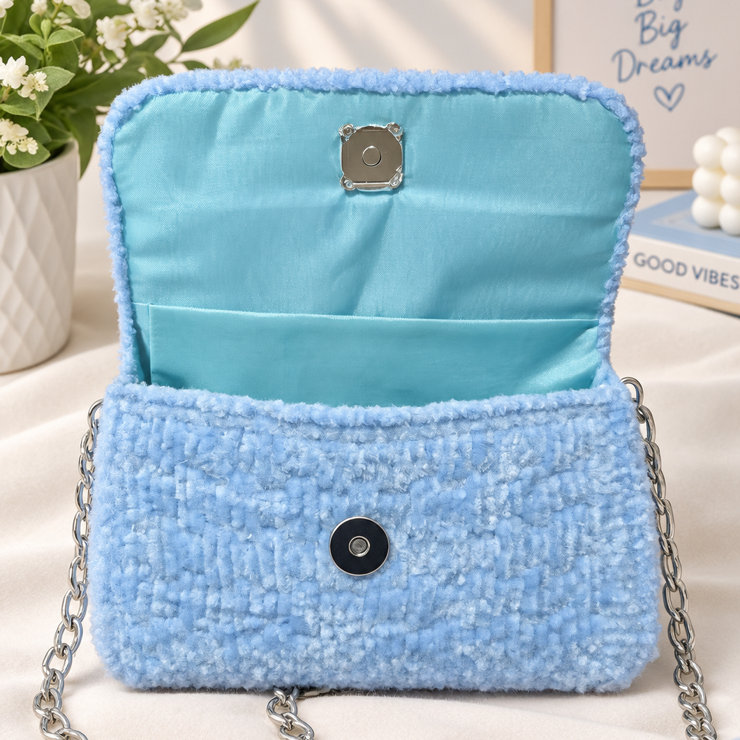 A light blue tweed shoulder bag with a silver chain strap, shown open to reveal a matching light blue satin lining. The bag is styled on a beige fabric surface with a potted plant, a book, and a decorative sign in the background.