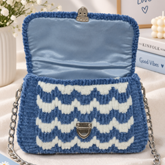 A blue and white chevron-patterned fuzzy handbag with a silver chain strap, styled on a cream fabric surface with decorative props including flowers, a candle, and books.