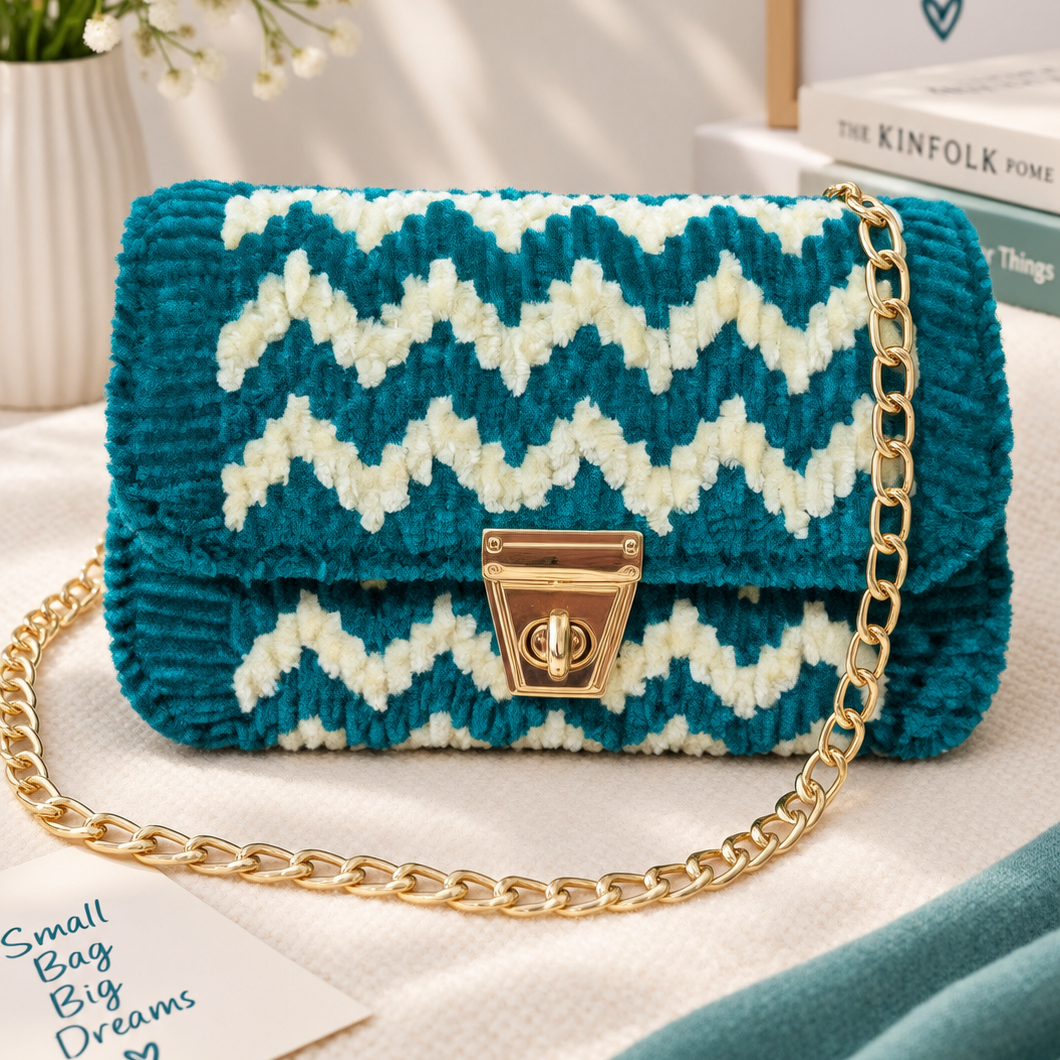 A small handbag with a teal and cream chevron patterned body and a gold chain strap, styled on a white fabric surface with decorative props including flowers, a sign, and a card.