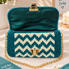 A teal and cream chevron-patterned knit purse with gold-tone hardware and a gold chain strap, styled on a light fabric surface with decorative flowers and a sign in the background.
