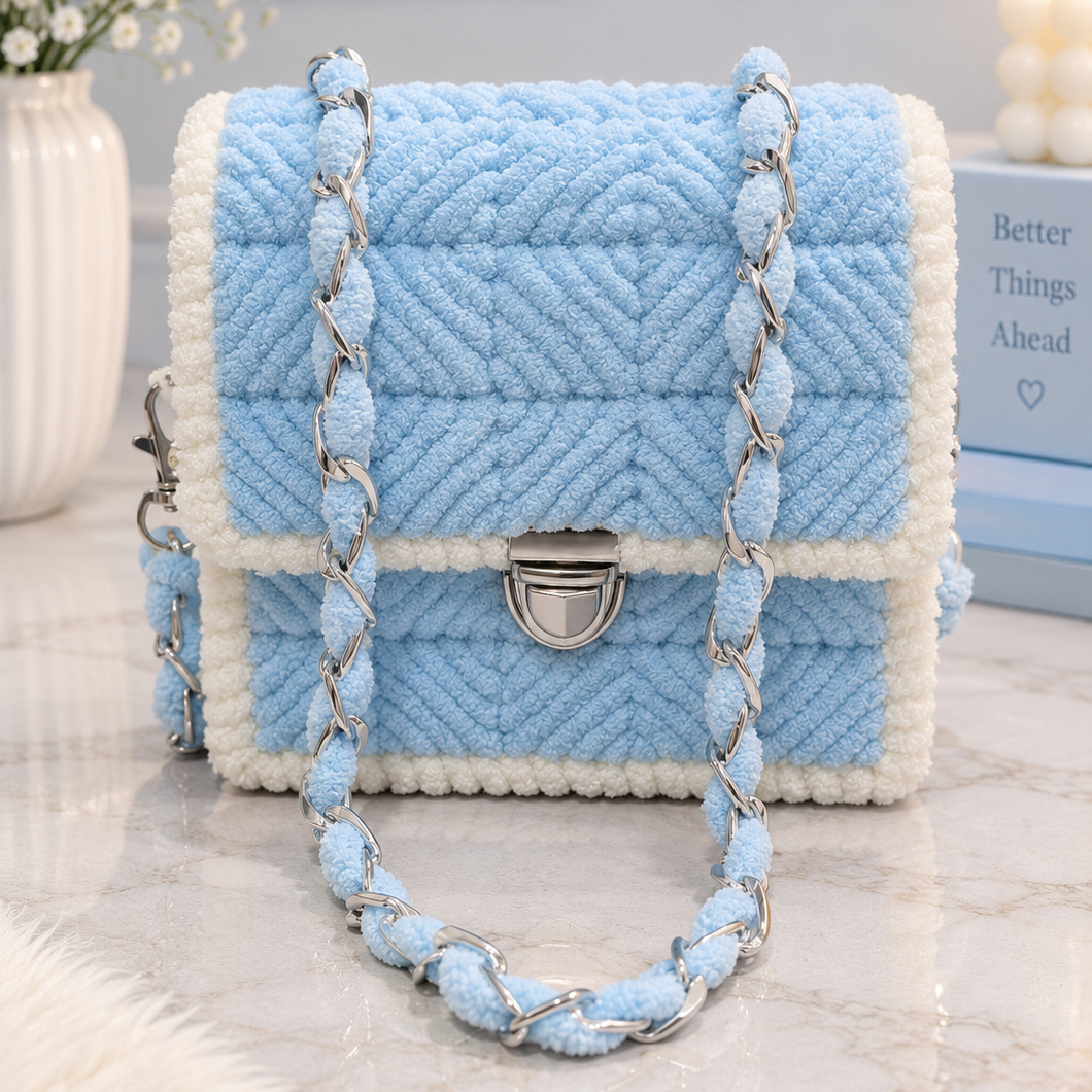A light blue and white textured handbag with a braided chain strap, displayed on a marble surface with decorative props in the background.