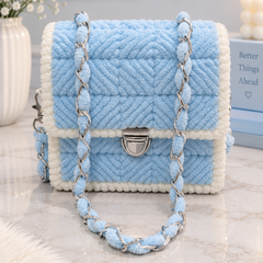 A light blue and white textured handbag with a braided chain strap, displayed on a marble surface with decorative props in the background.