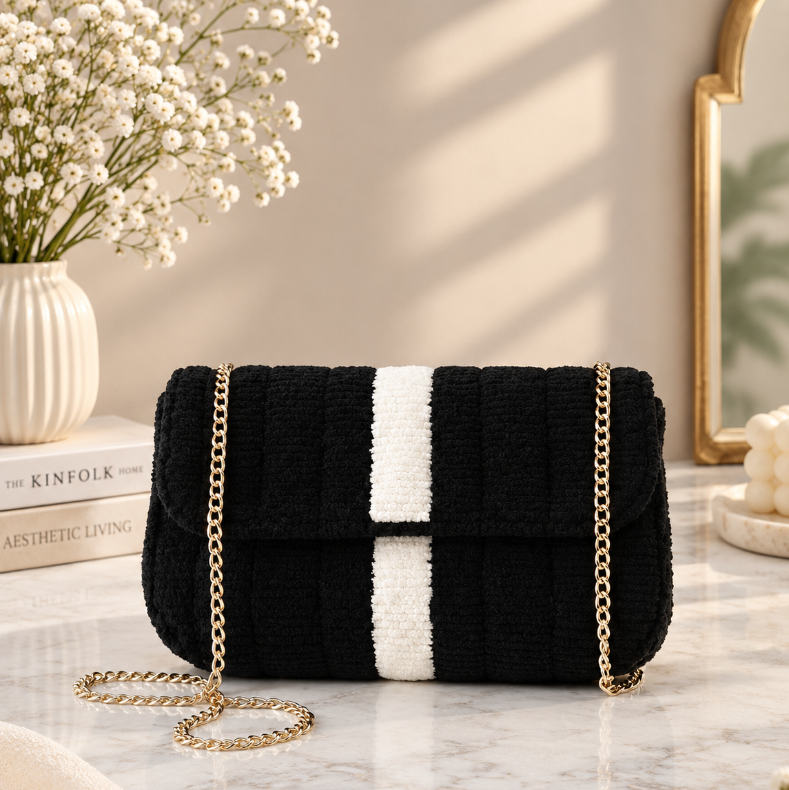 A black and white striped textured shoulder bag with a gold chain strap, styled on a marble tabletop with a vase of baby's breath and a book in the background.