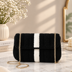 A black and white striped textured shoulder bag with a gold chain strap, styled on a marble tabletop with a vase of baby's breath and a book in the background.