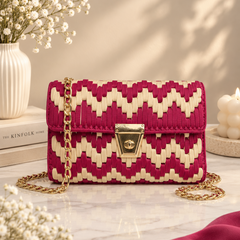 A woven clutch handbag with a magenta and cream chevron pattern and a gold chain strap, styled on a marble surface with decorative flowers and candles.