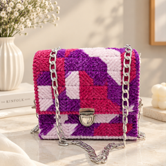 A small structured handbag with a chunky woven geometric pattern in purple, pink, and white, featuring a silver chain strap and a silver turn-lock clasp, displayed on a marble surface with a vase of flowers in the background.