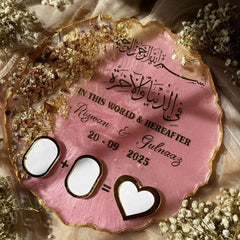 Personalized pink plaque with Arabic text and dates, surrounded by flowers on a soft fabric background.